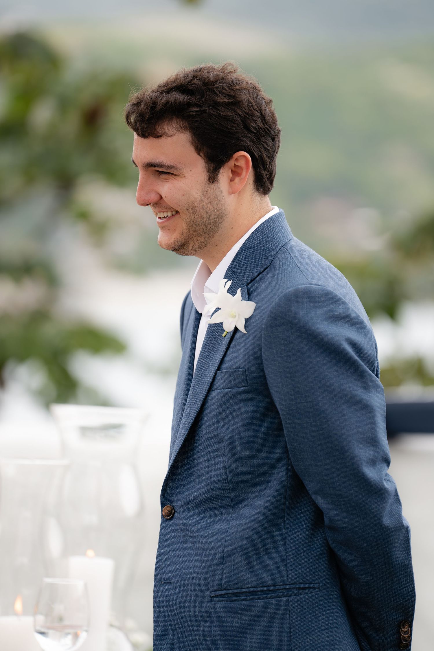 Groom in blue suit smiles during outdoor wedding ceremony.