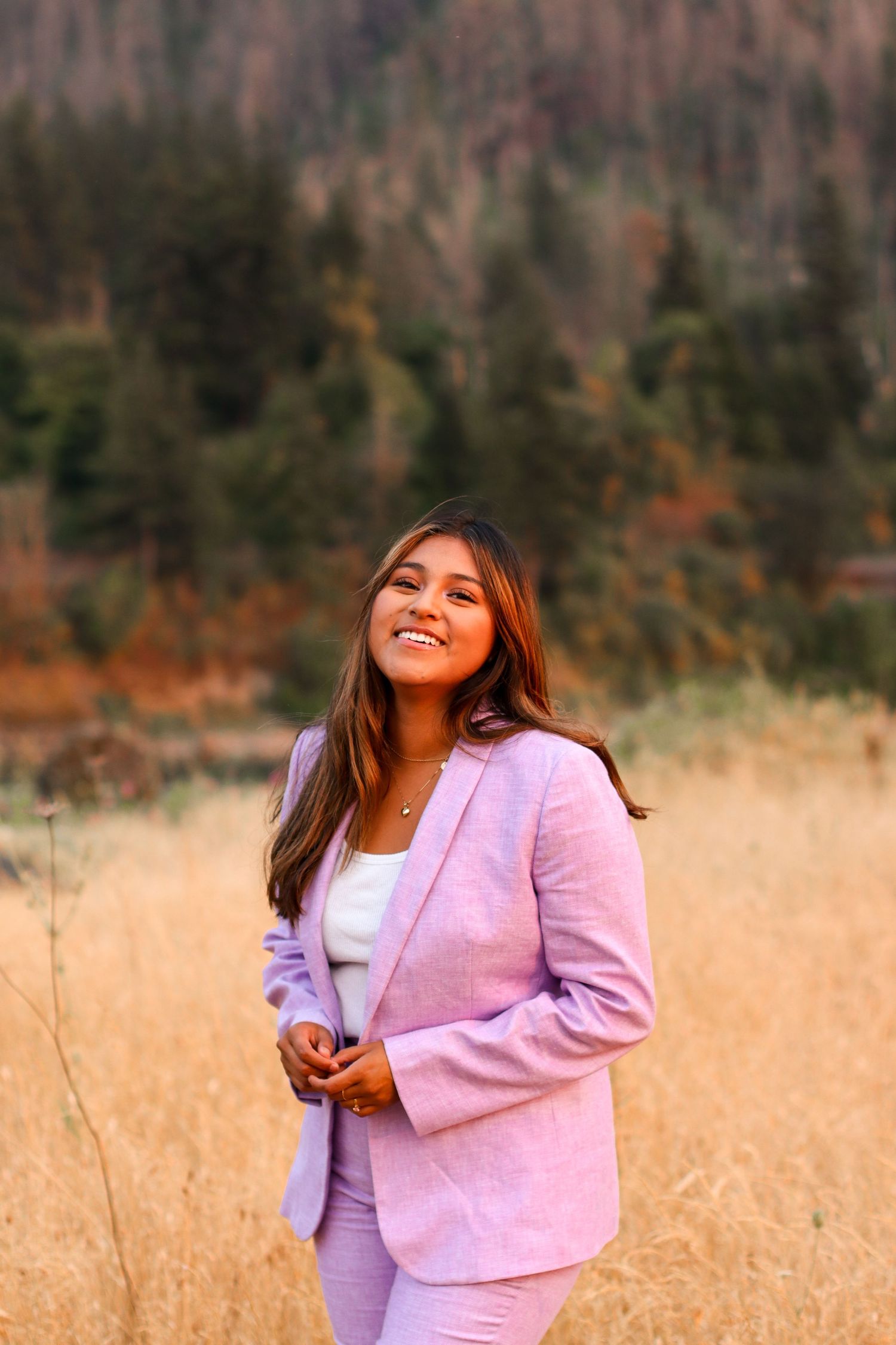 Person in pink blazer poses in autumn field with mountains in background.
