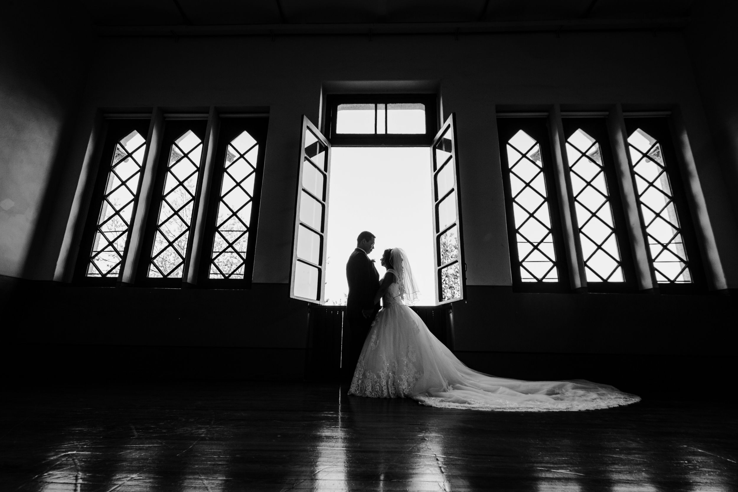 Dramatic silhouette of newlyweds against Gothic windows in a dark, elegant interior space.