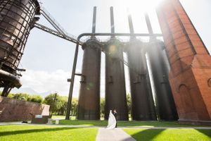 Wedding photo taken near industrial architecture with tall metal structures and sunlight.