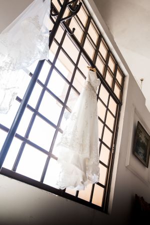 Wedding dress hangs dramatically in front of large geometric window with natural lighting.