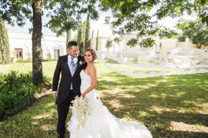 Couple poses in formal wedding attire on sunny lawn with classic architecture in background.