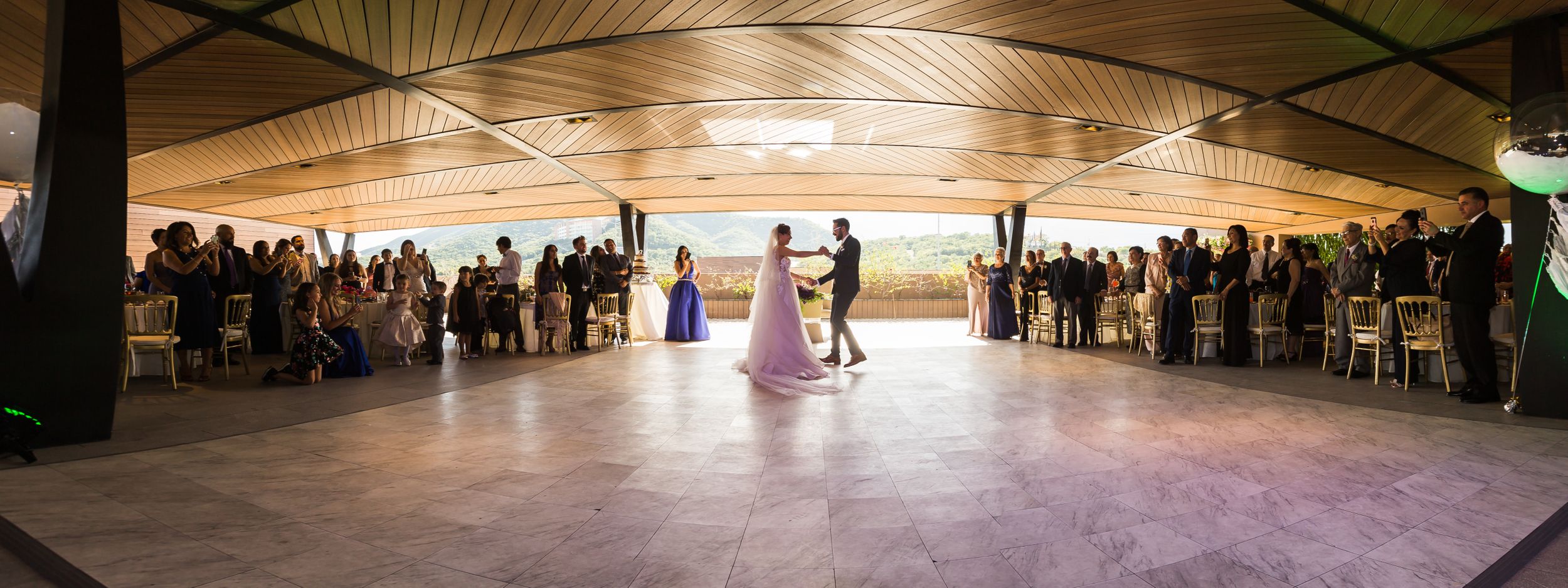Panoramic view of wedding guests gathered around an empty dance floor under a wooden pavilion ceiling.