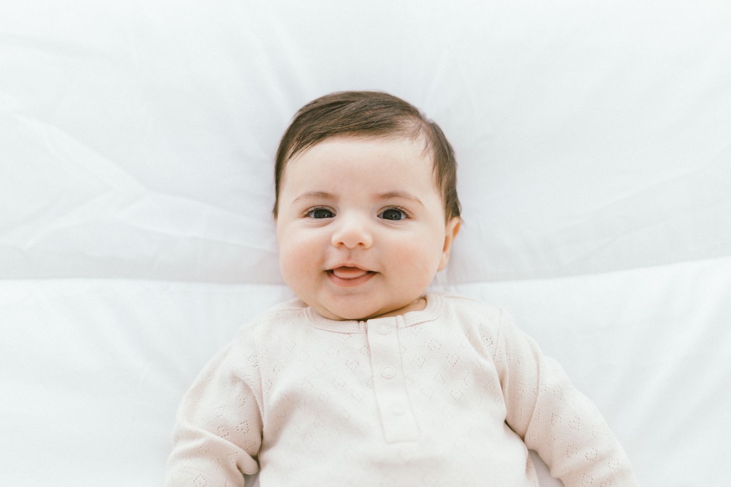 Close-up of smiling baby in beige outfit lying on white bedding.