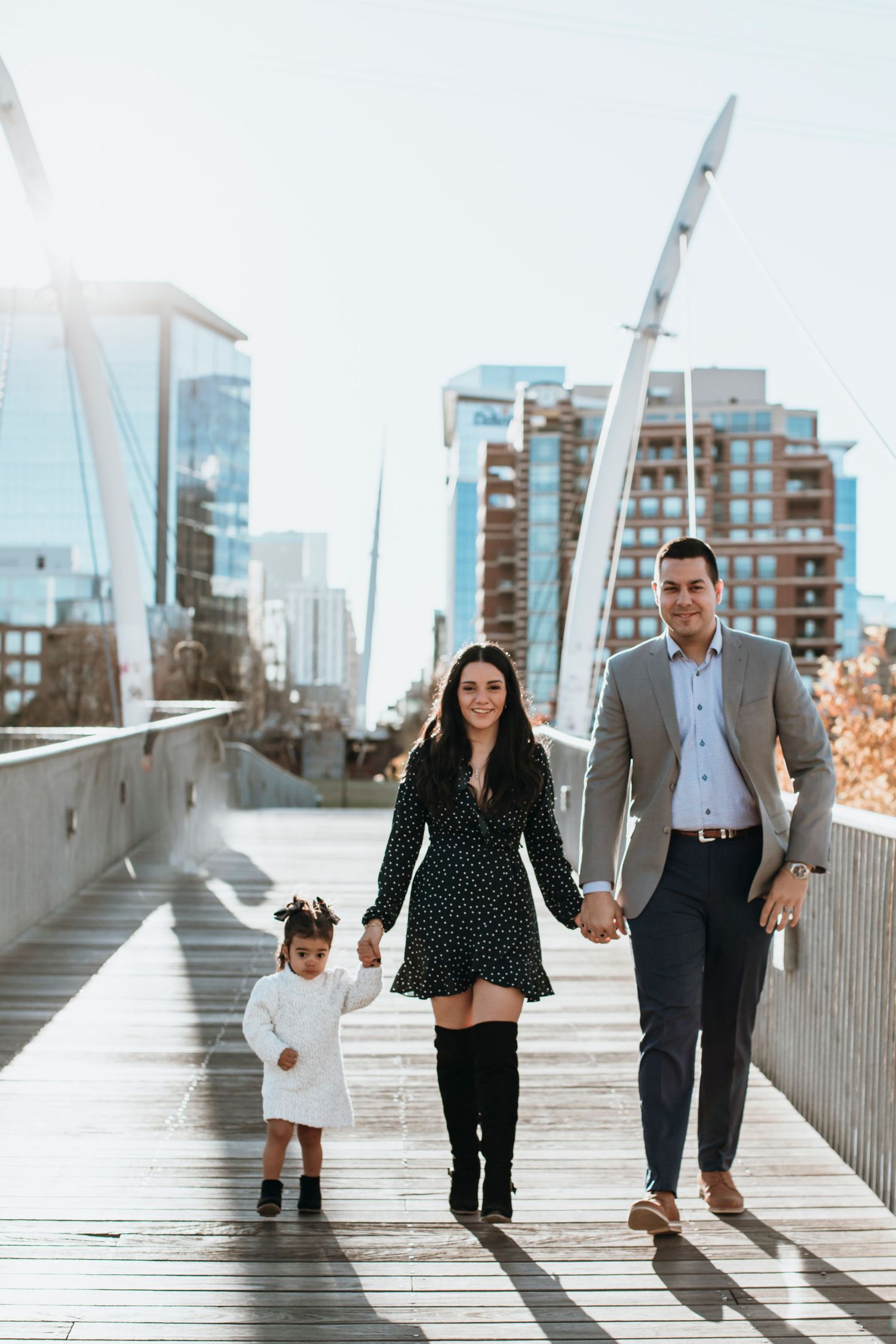 A family of three walking on a bridge and smiling at the camera with the sun shining behind them
