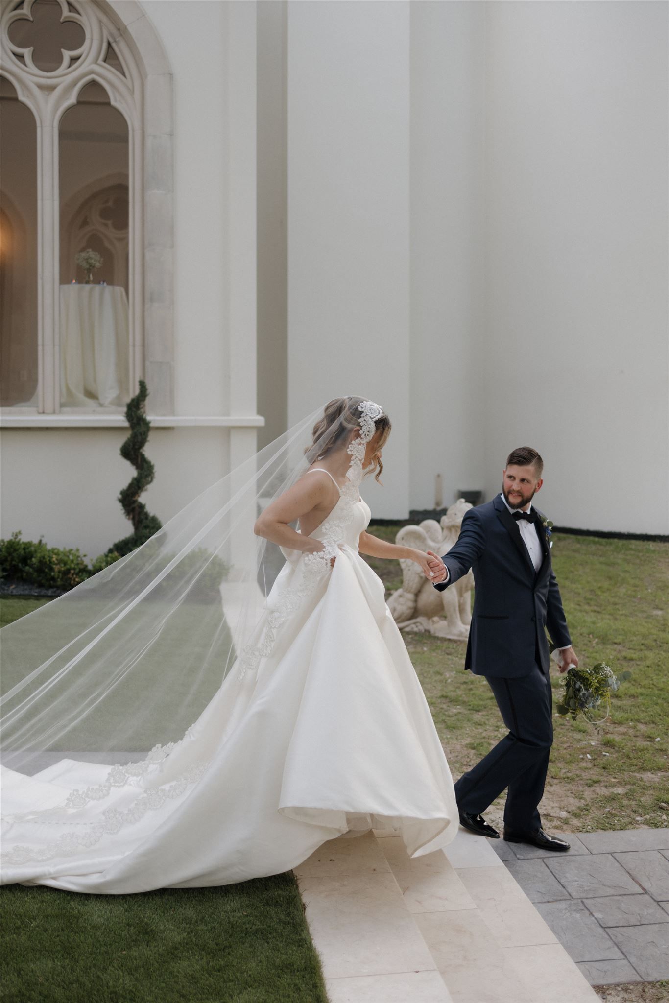A flowing wedding veil trails behind couple walking outside gothic-style church architecture.