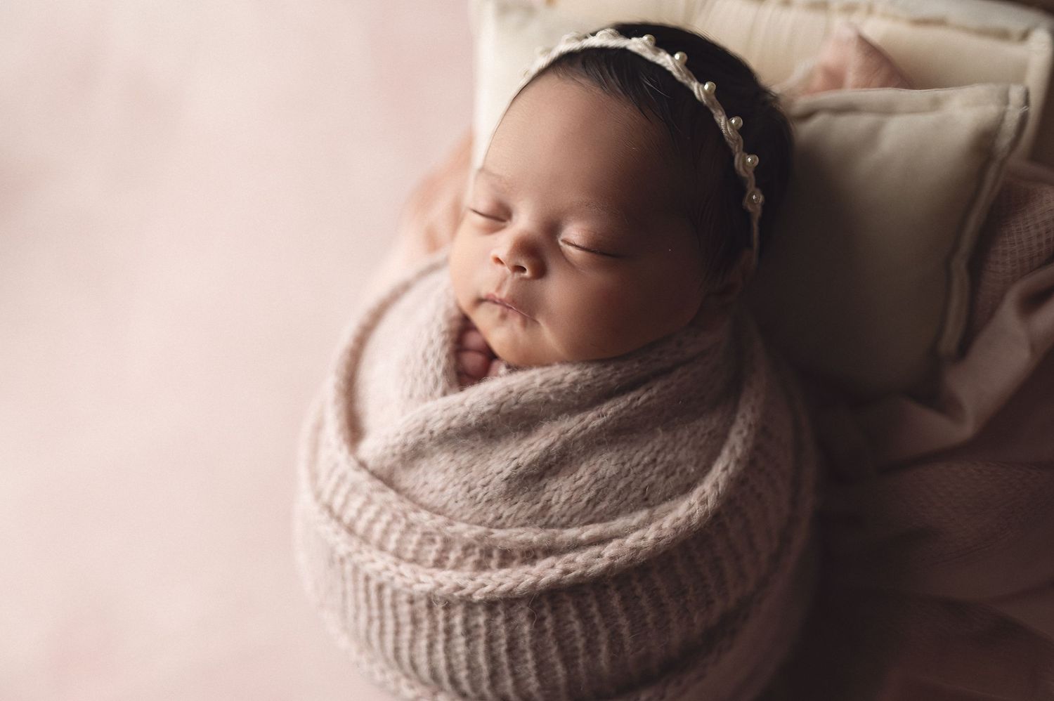 Cozy newborn wearing delicate headband sleeps wrapped in knitted swaddle.