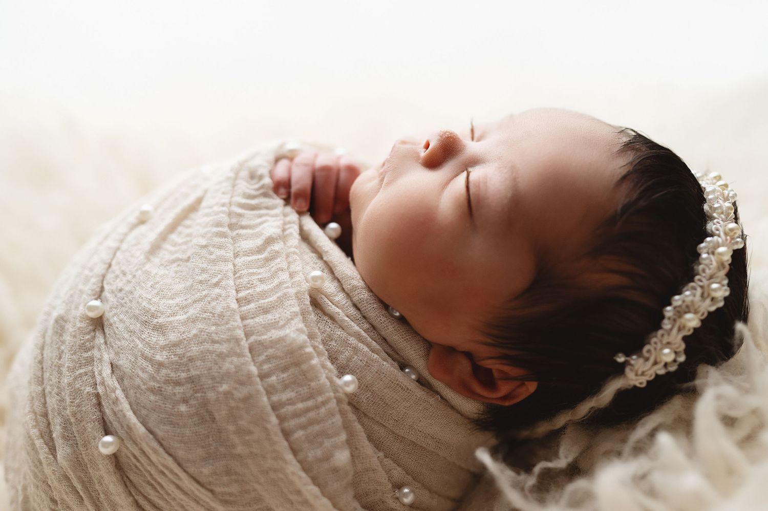 Close-up of sleeping infant with delicate headband swaddled in white fabric.
