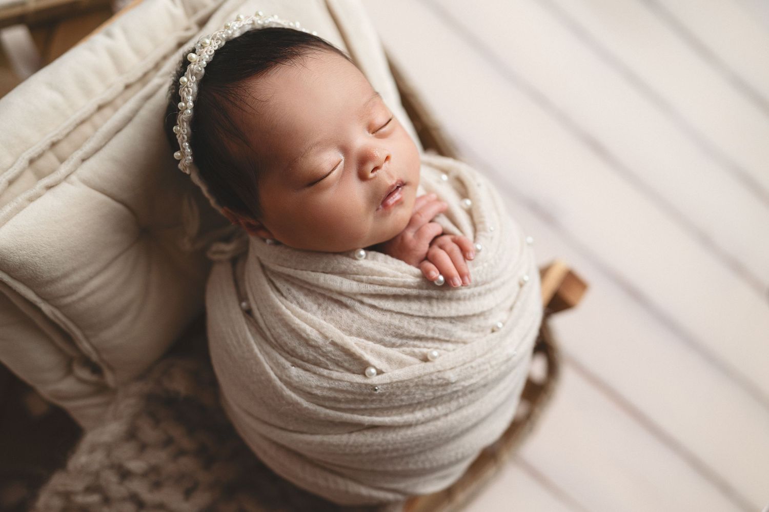 Sleeping newborn swaddled in white fabric rests on textured cream cushion.