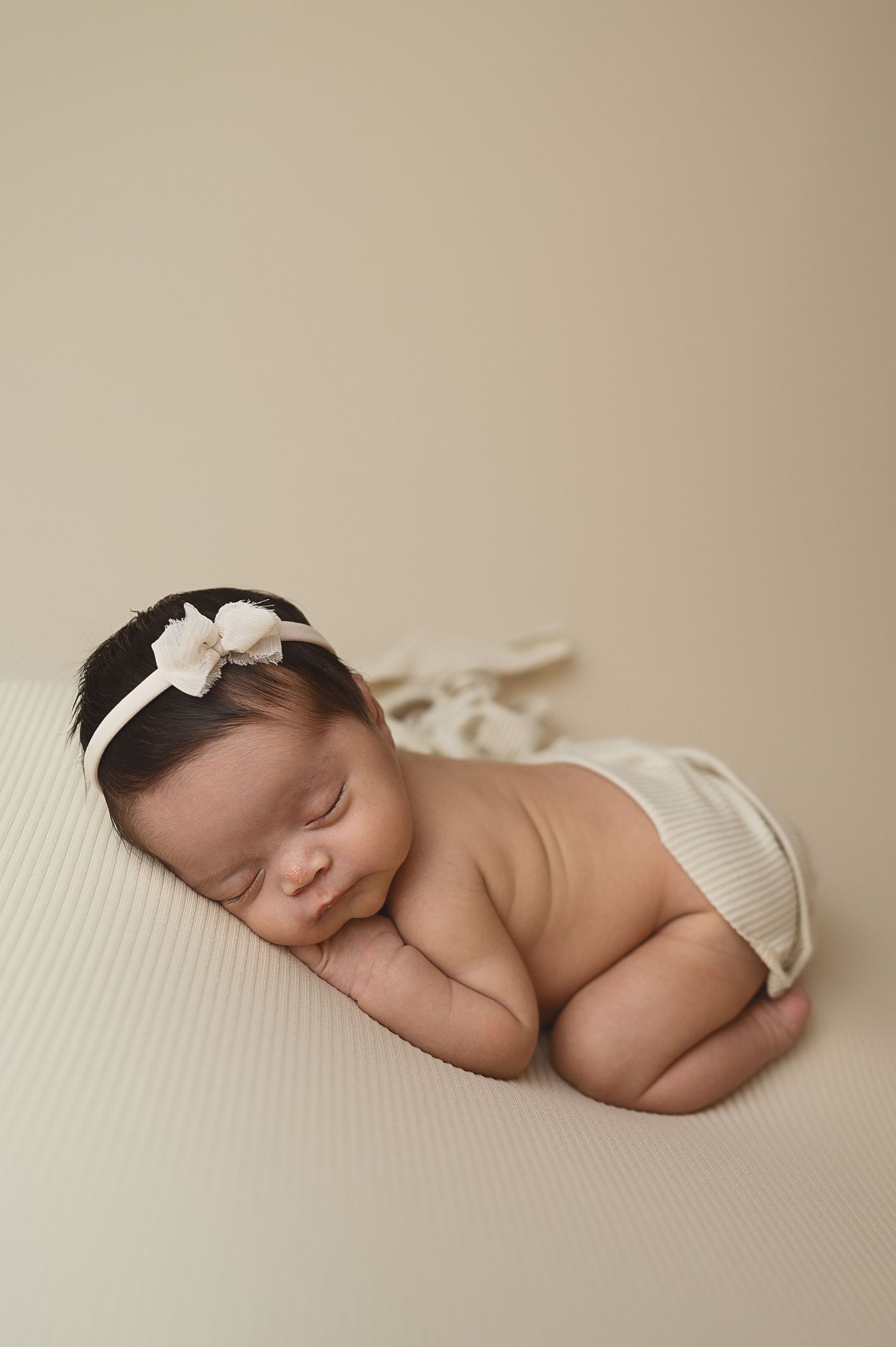 Sleeping newborn with white bow headband posed on cream backdrop.