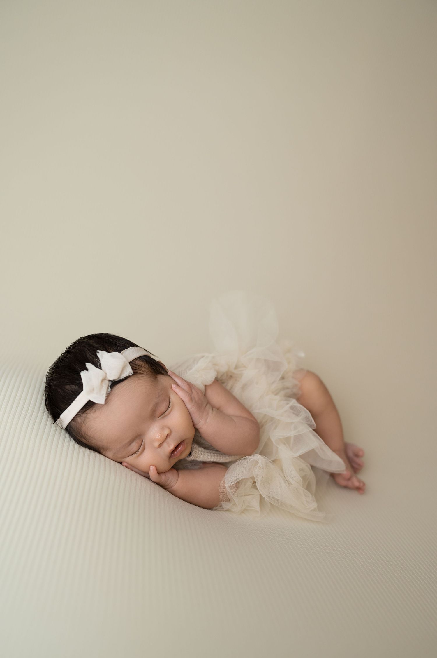 Sleeping newborn with white bow headband draped in soft cream fabric.