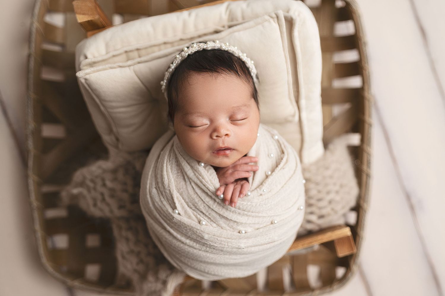Newborn baby wrapped in soft white swaddle sleeps peacefully in wooden basket on cream pillow.