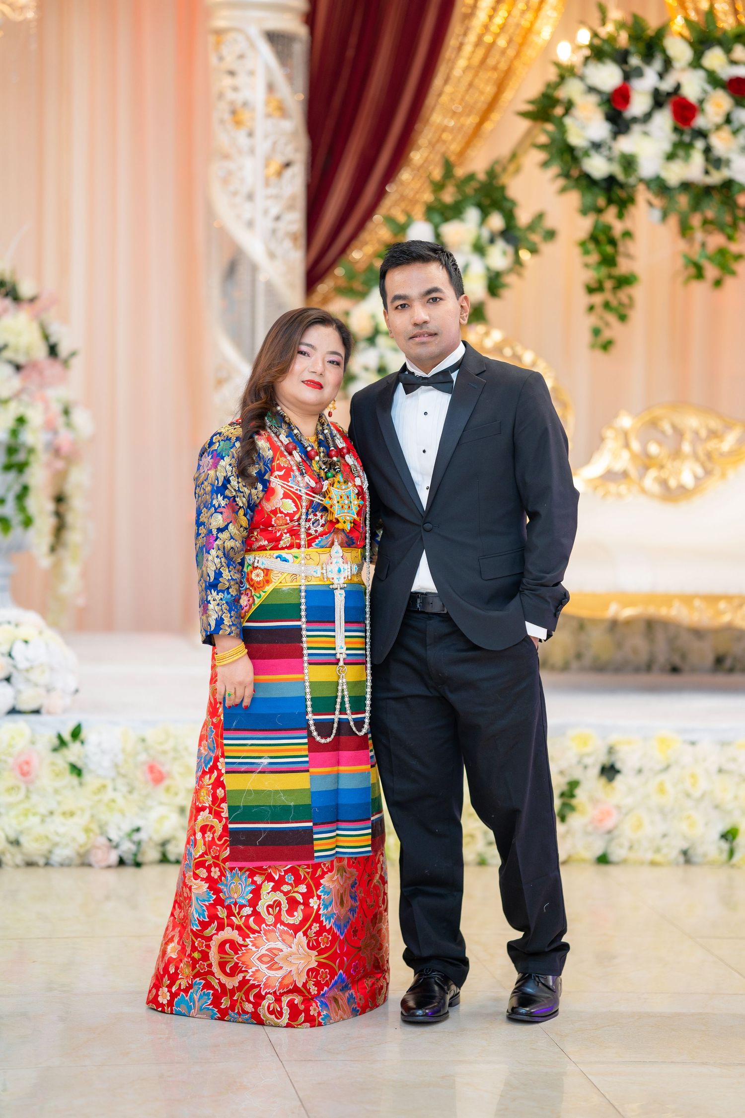 A couple poses elegantly against a floral backdrop wearing formal attire.