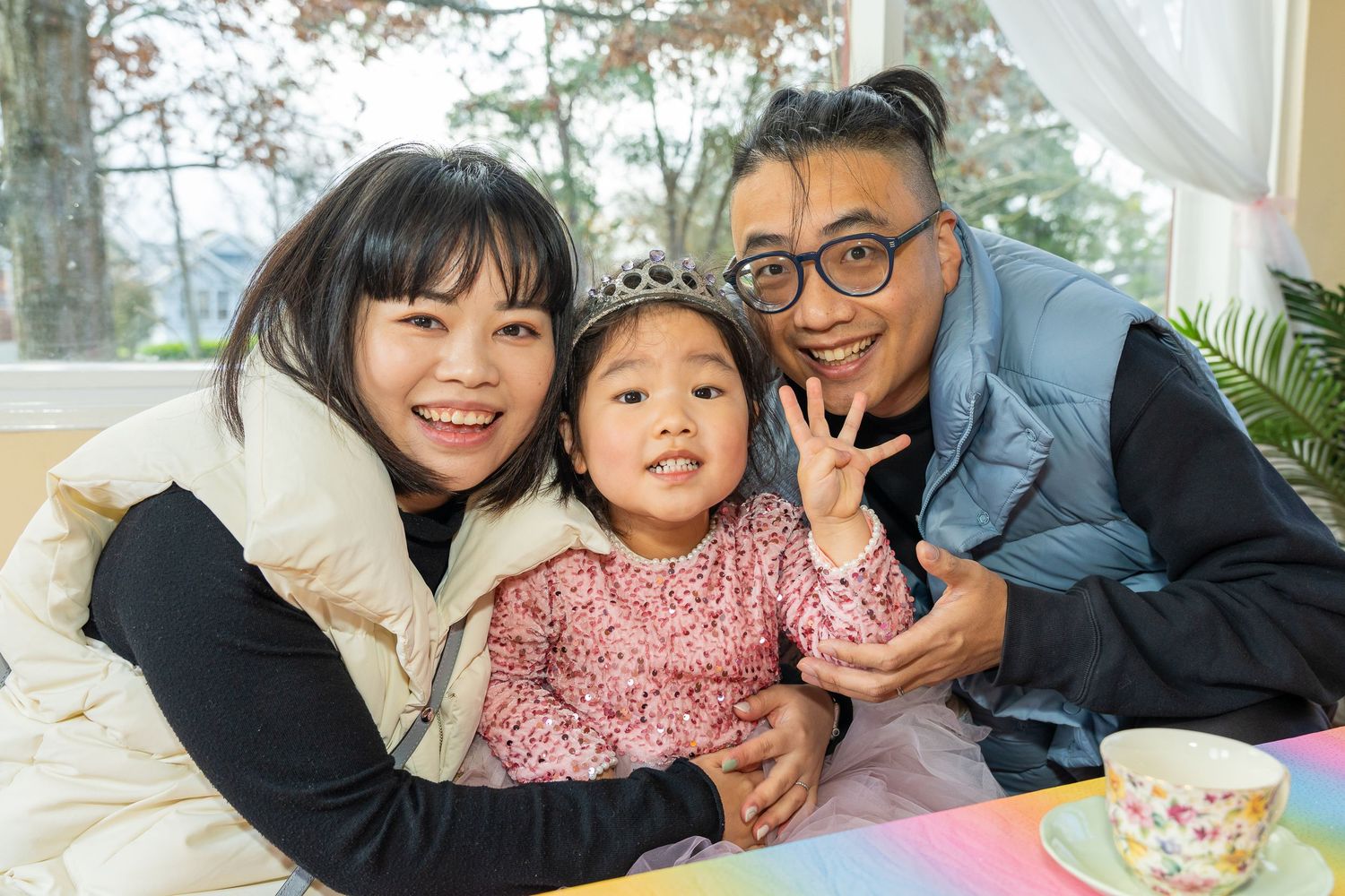 Smiling Asian family of three posing closely together at a colorful princess tea party table.