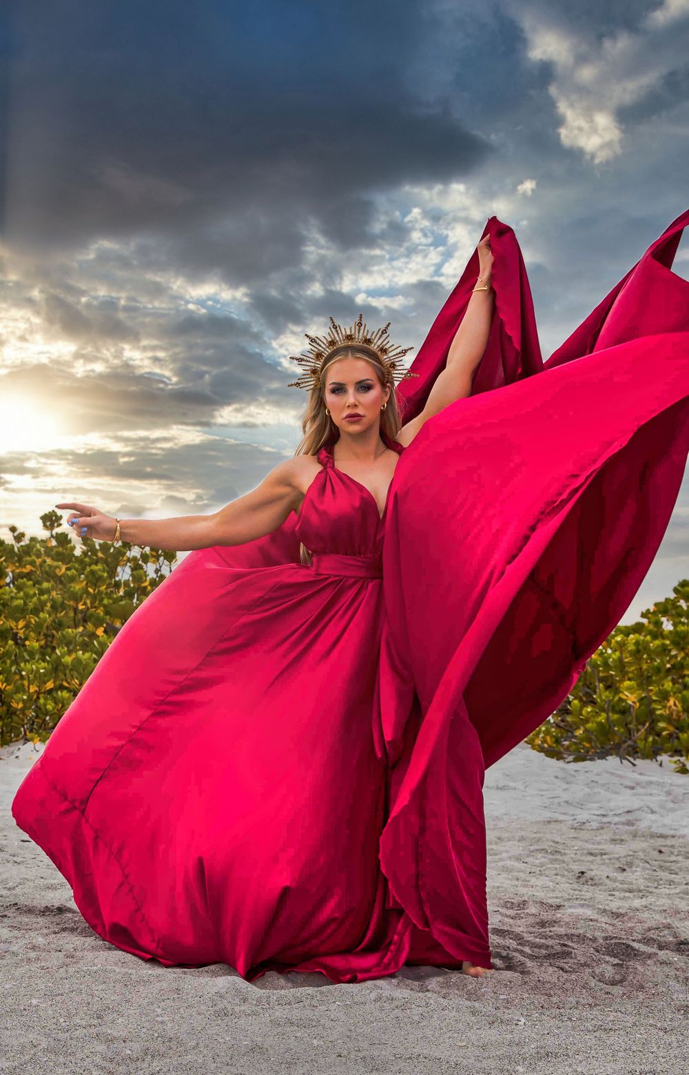 Dramatic red dress catches wind against sunset sky and foliage.
