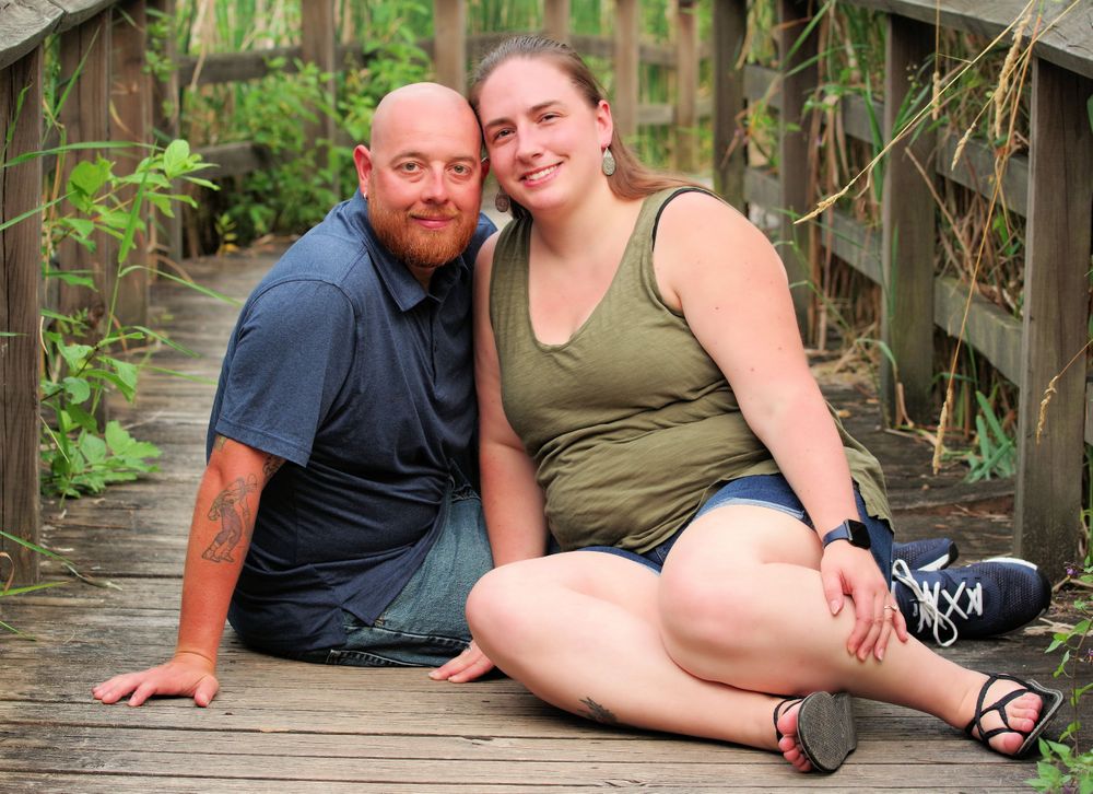 A couple sits casually together on a wooden boardwalk path through nature.