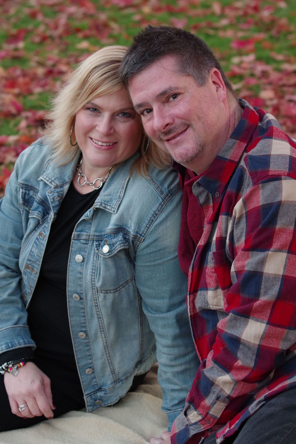 Couple in casual denim and plaid posing together against fall foliage background.