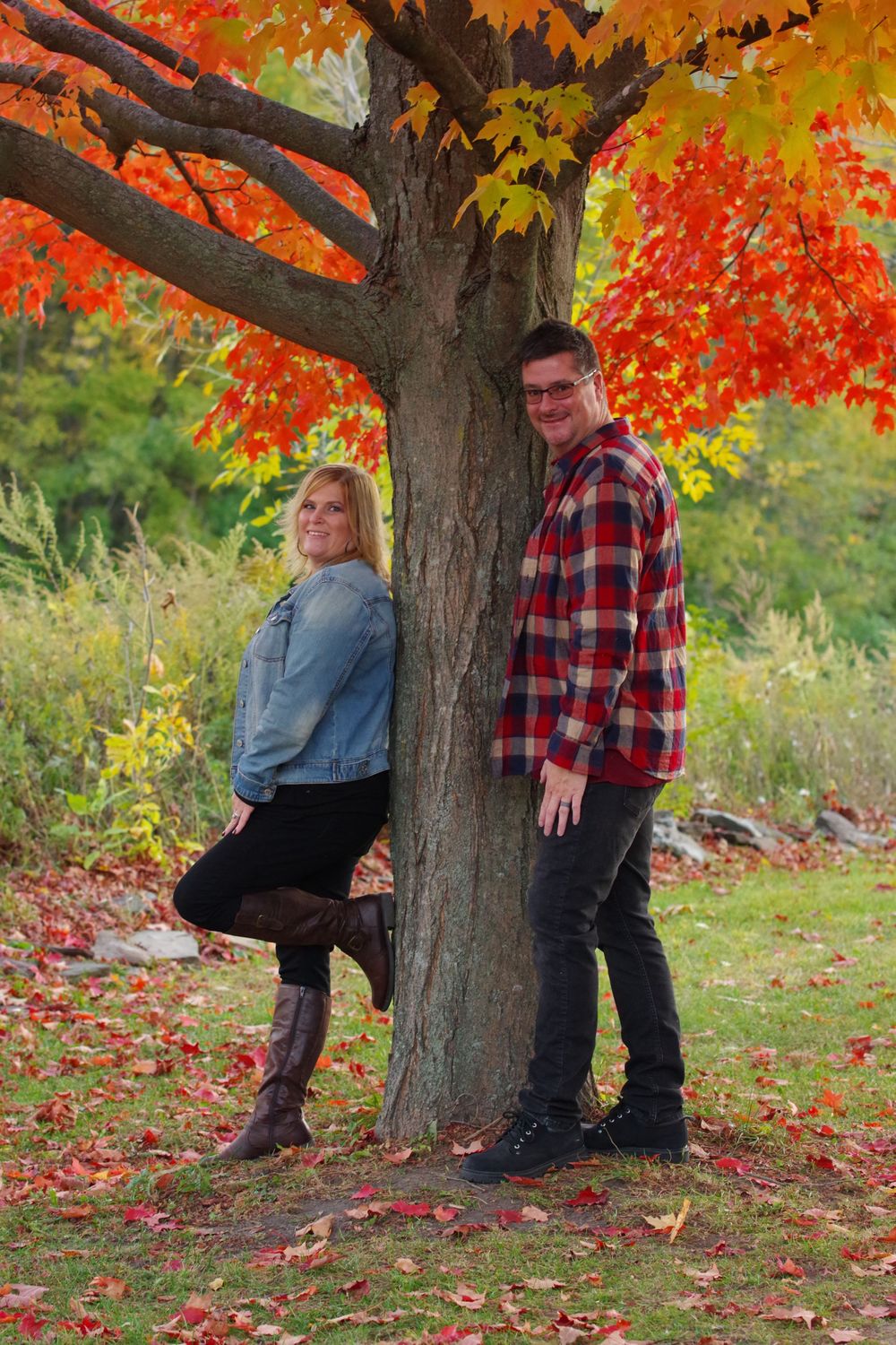 Couple poses playfully against tree trunk with bright orange autumn leaves overhead.