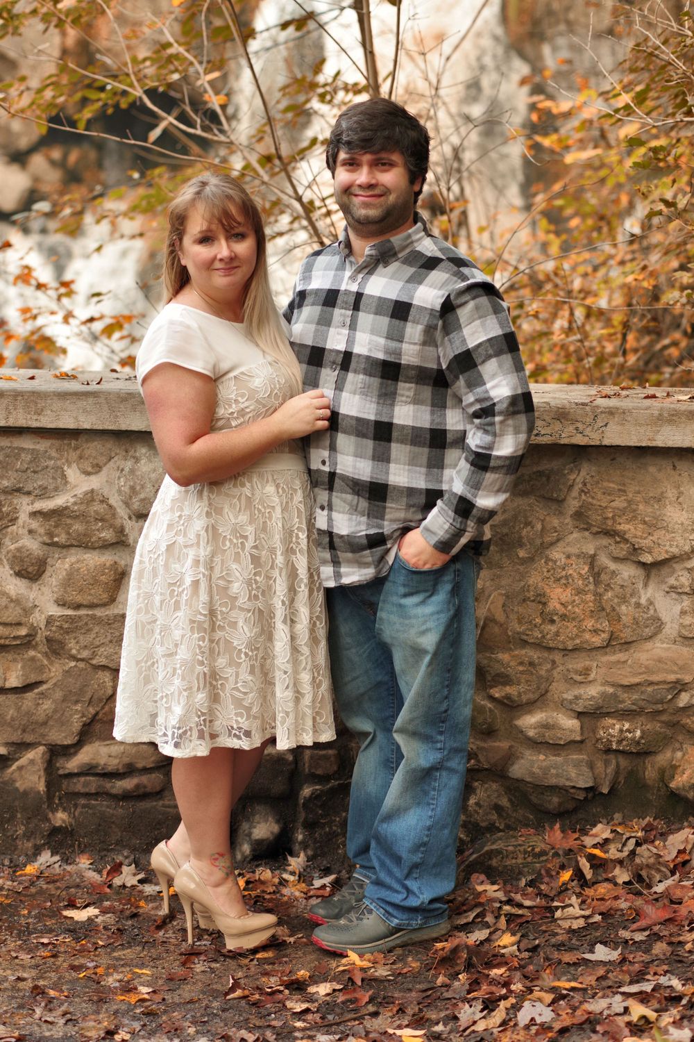 A couple poses against a stone wall surrounded by fall foliage.
