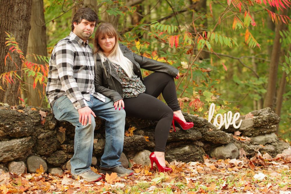 A couple sits on a log surrounded by fall foliage with a decorative love sign.