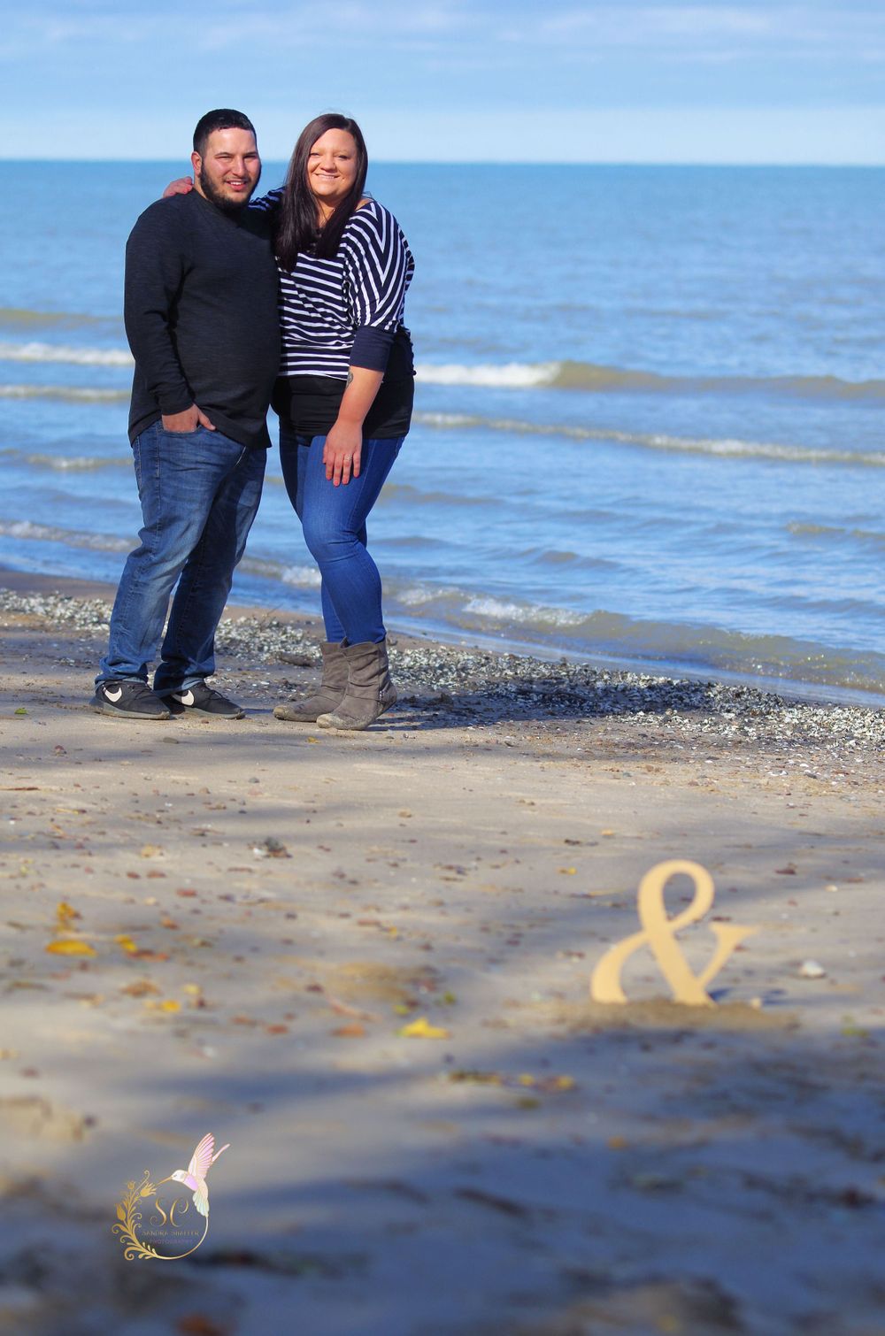 Couple stands on beach with ampersand symbol drawn in sand at their feet.