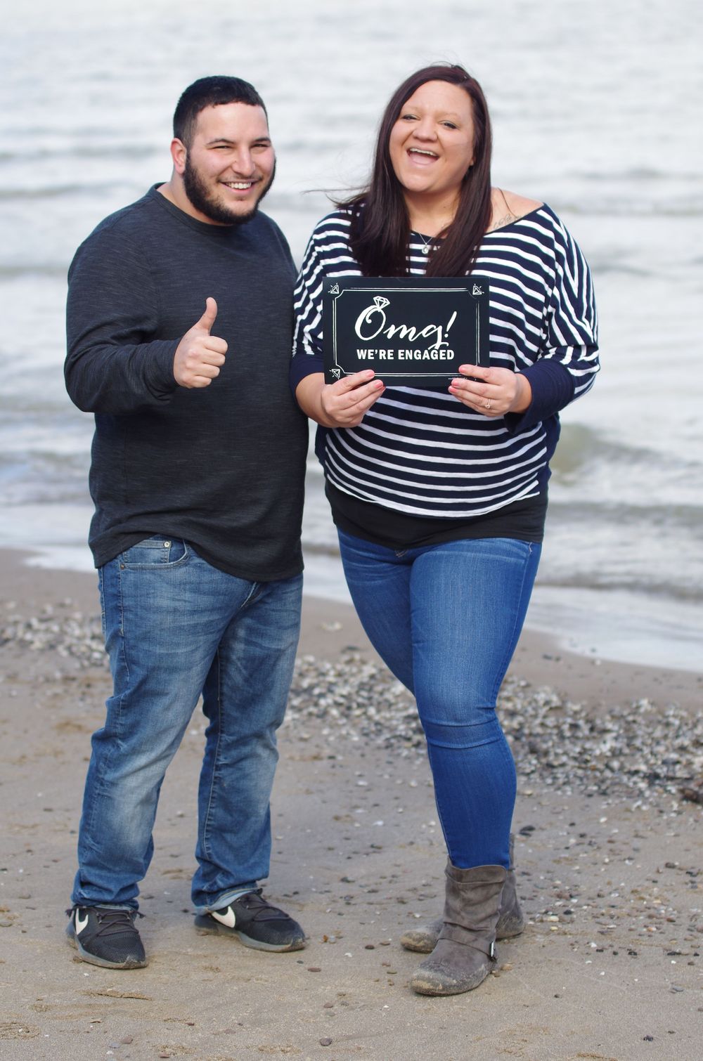 Couple on beach holding a sign and giving thumbs up during pregnancy announcement photo shoot.