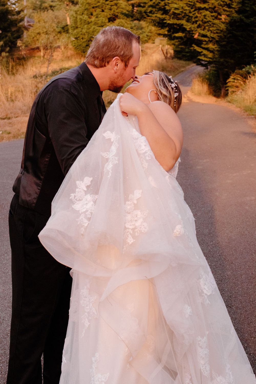 Newlywed couple shares romantic kiss on a sunlit road, bride in lace gown with floral veil flowing in breeze.