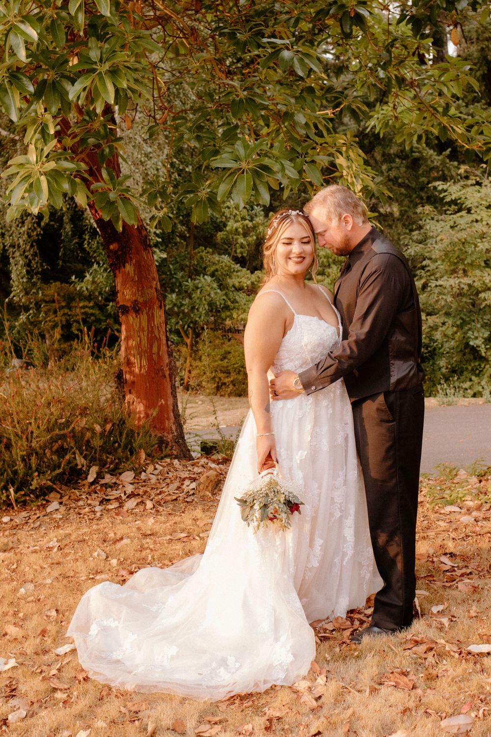 Bride in lace wedding gown holding bouquet while groom embraces her under autumn trees.