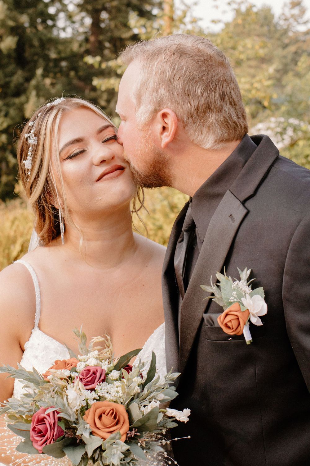 Groom in black suit kissing bride on cheek while she holds colorful rose bouquet outdoors.