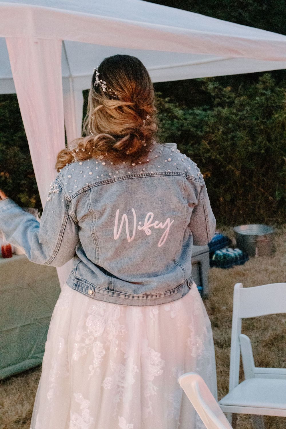 Bride wearing a pearl-embellished denim jacket with Wifey script at an outdoor wedding reception.