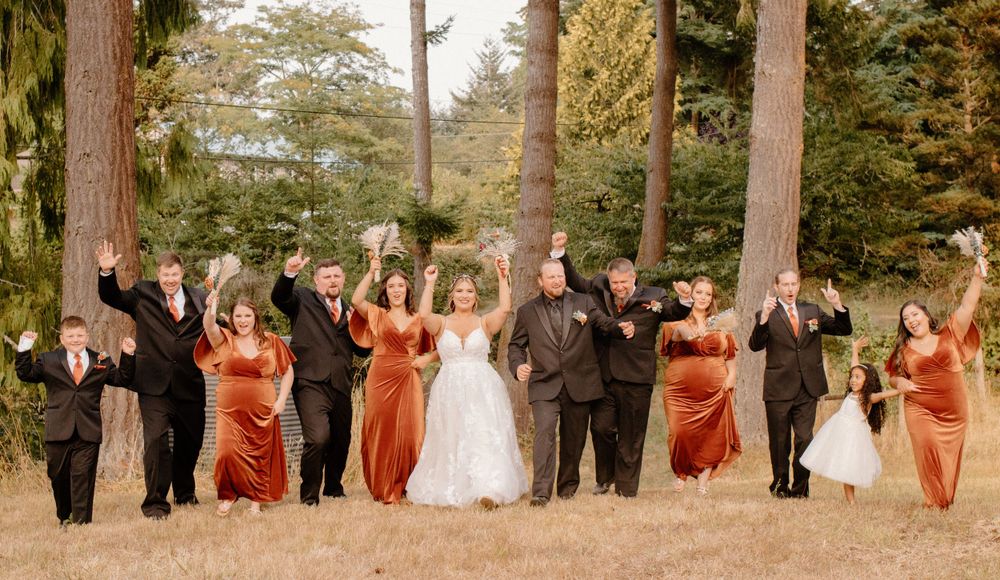 Wedding party in black and rust velvet dresses celebrating with raised arms among tall trees.