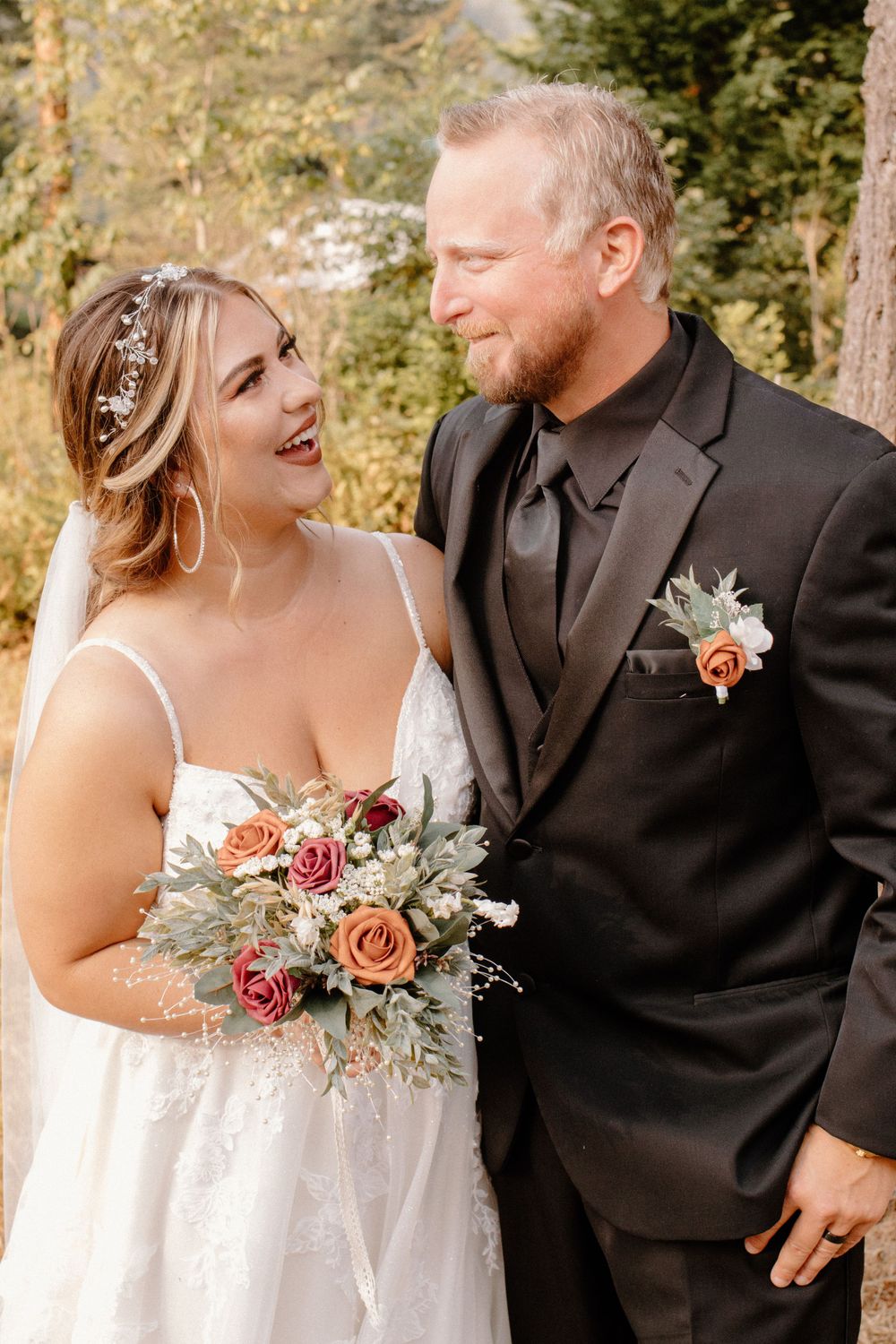 Bride and groom laughing together outdoors, holding rose bouquet, groom wearing black suit.