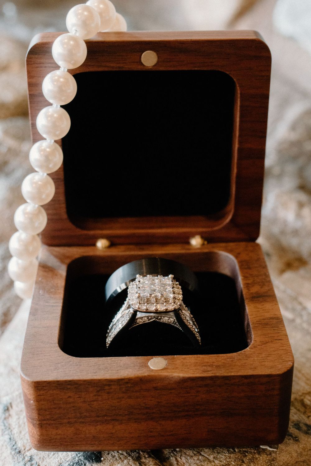 Diamond halo engagement ring displayed in an open wooden ring box with pearl necklace.