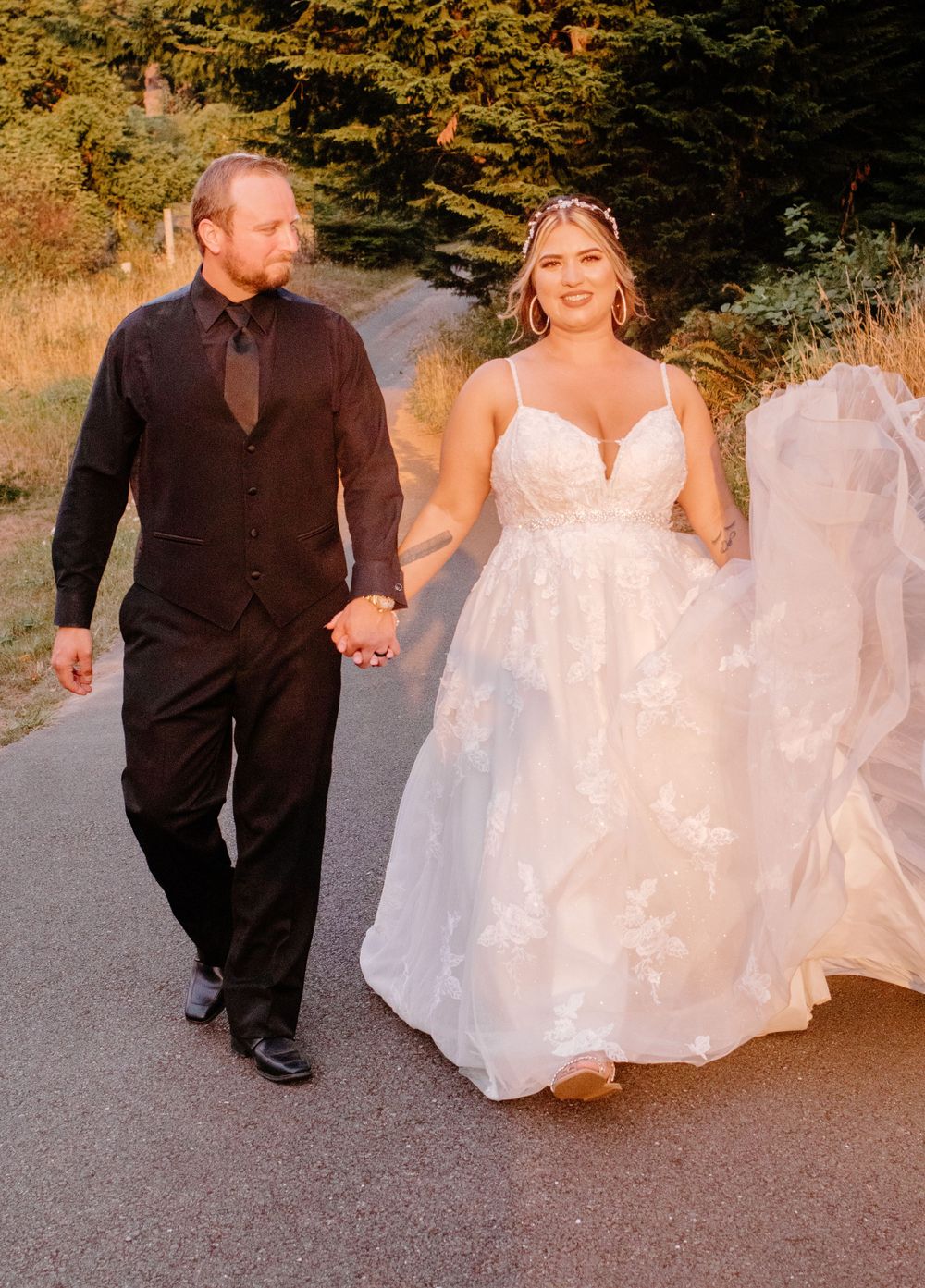 Bride and groom walk hand in hand down a tree-lined path, bride wearing lace wedding gown with flowing veil.