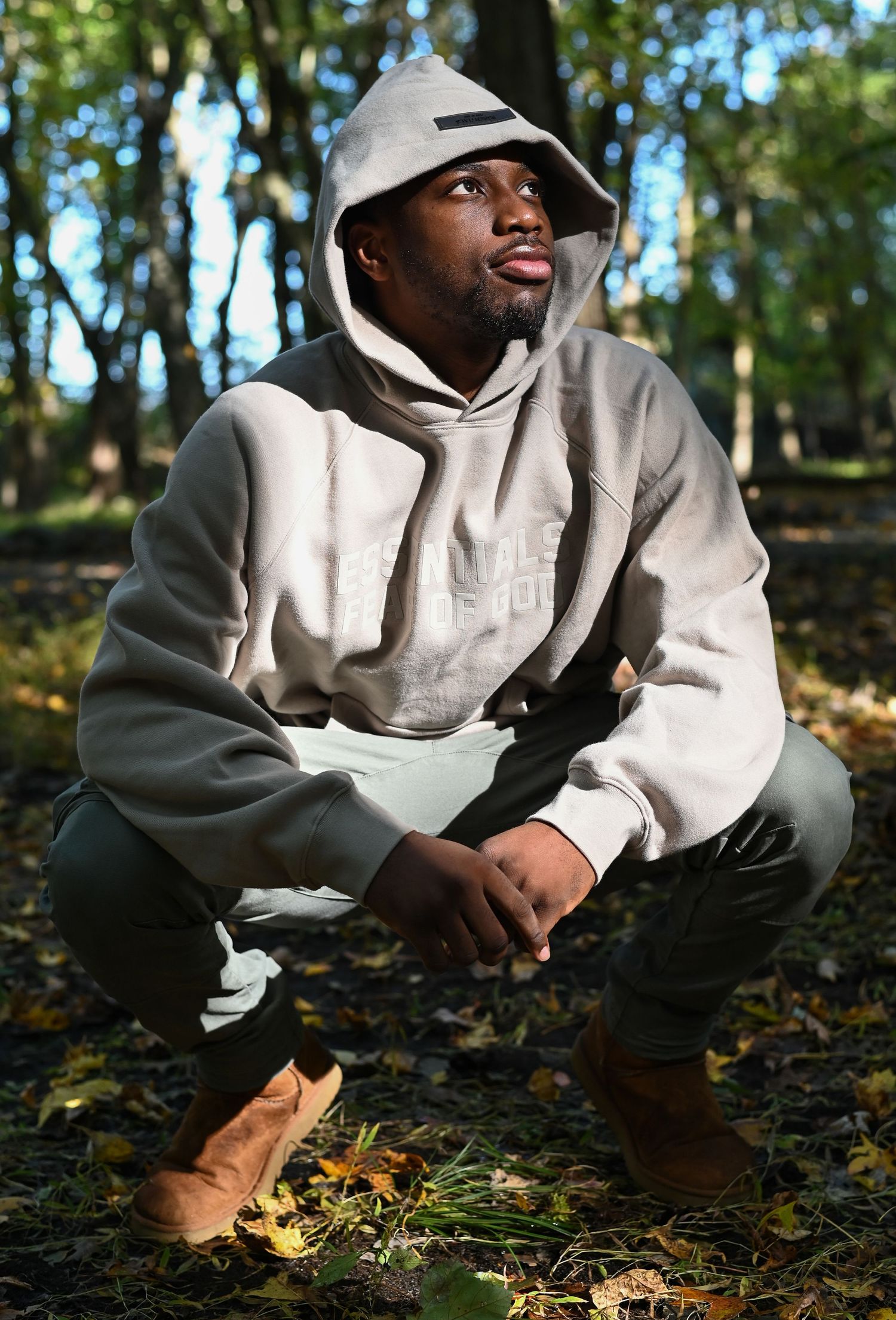 Someone in a light gray hoodie crouches among autumn leaves in a wooded setting.