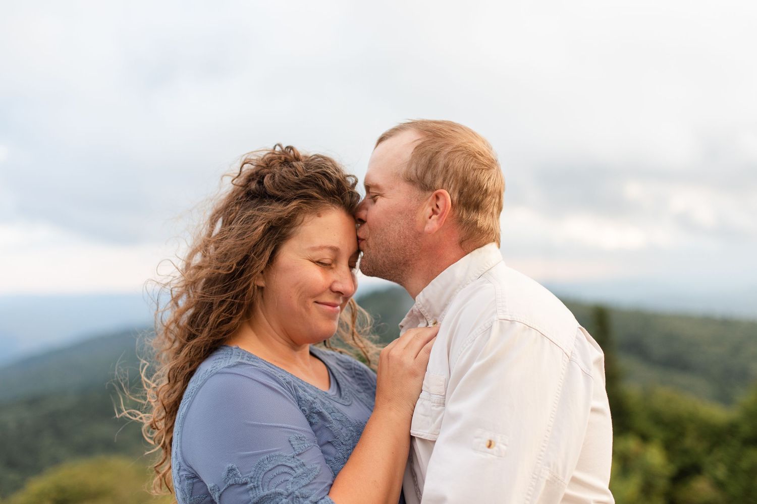 Romantic couple embraces on a mountaintop overlooking scenic mountain vistas during sunset.