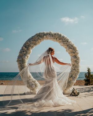 White floral wedding arch by the ocean with flowing bridal veil catching the breeze.