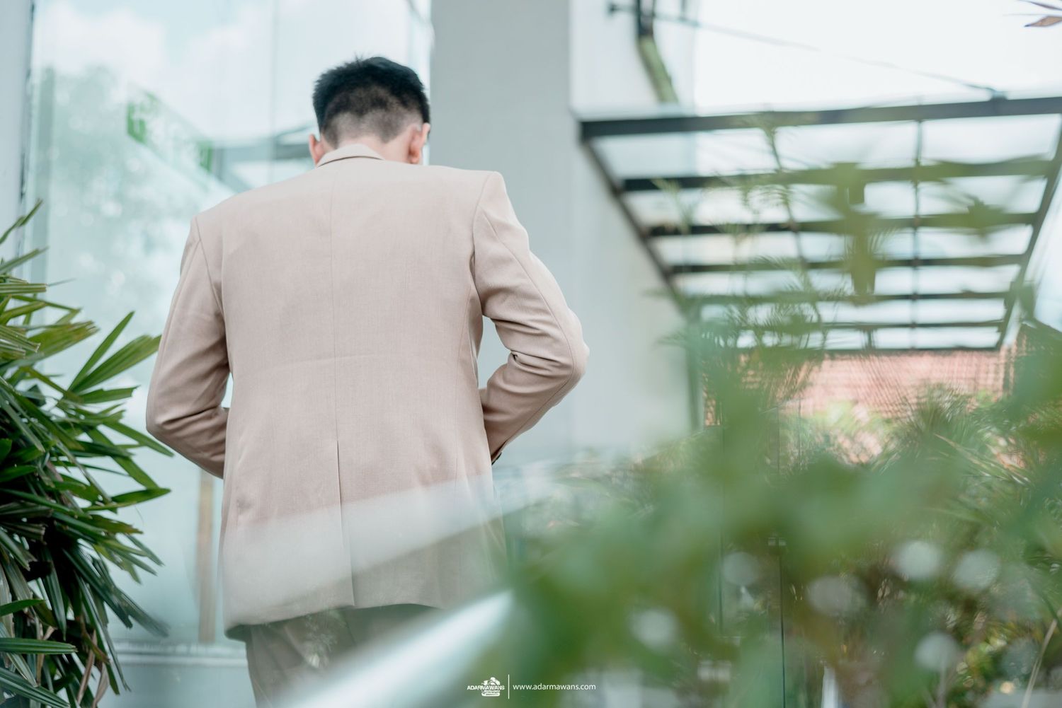 Man in beige suit jacket standing on balcony surrounded by green plants.