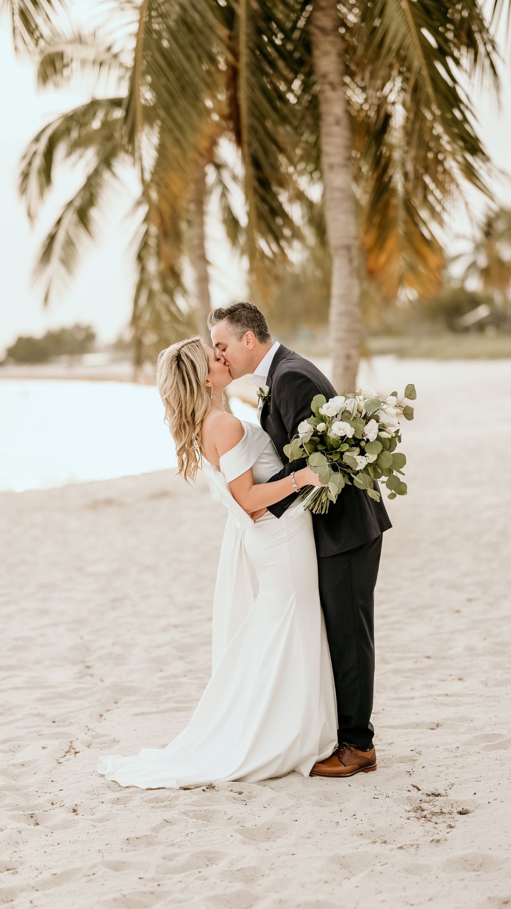 Romantic beach wedding portrait with palm trees and white floral bouquet.