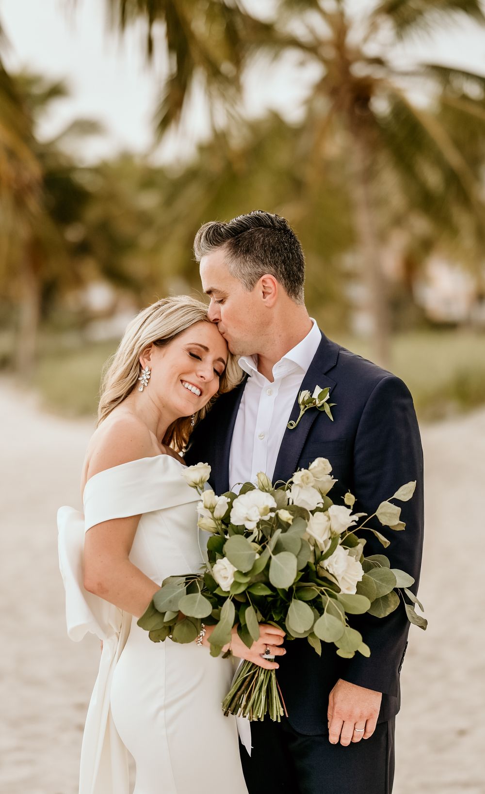 Intimate beach wedding moment with white and green bouquet against palm trees.