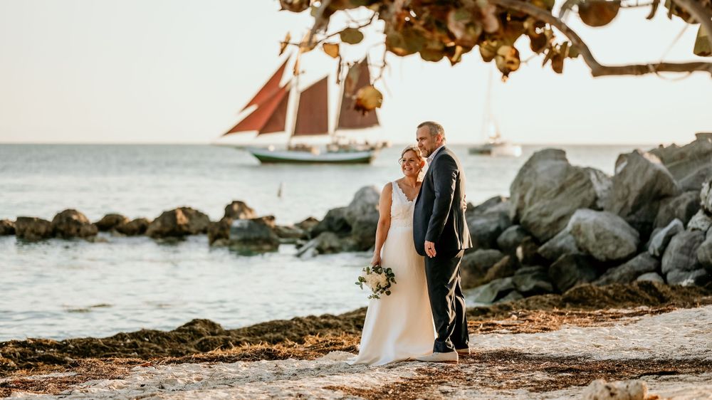 A couple poses for wedding photos on a beach with a sailboat visible in the background.