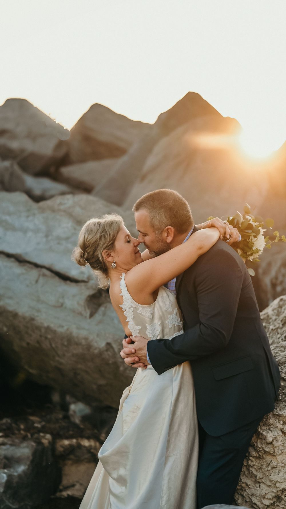 A bride and groom share a romantic kiss among rocky cliffs at sunset.