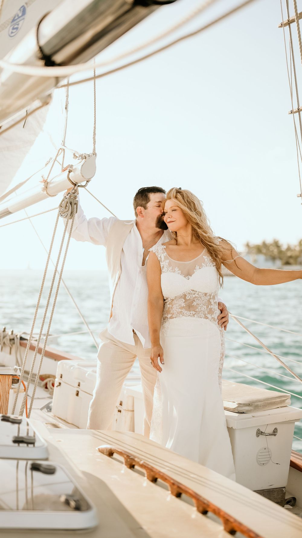 Wedding couple in white attire standing together on sailboat deck.