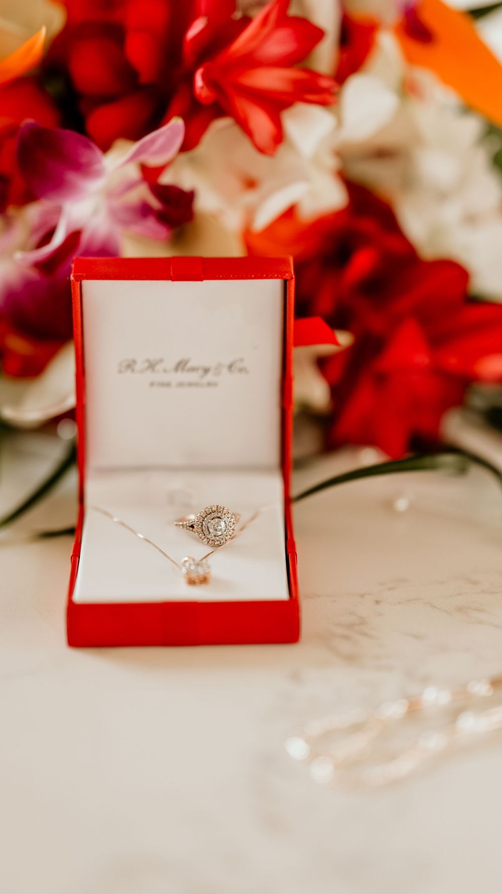 Delicate engagement ring and necklace in red box surrounded by vibrant flowers.