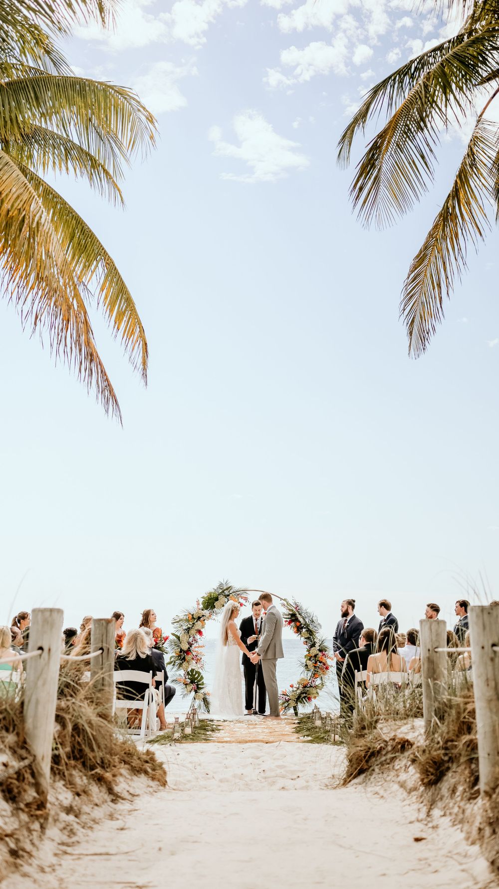 Beach wedding ceremony setup with palm trees framing a sandy path leading to an arch by the ocean.