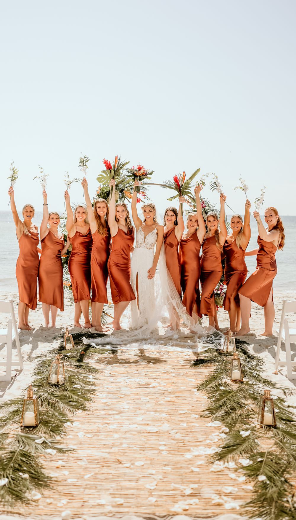 Bridal party in rust-colored dresses celebrating on beach wedding aisle.