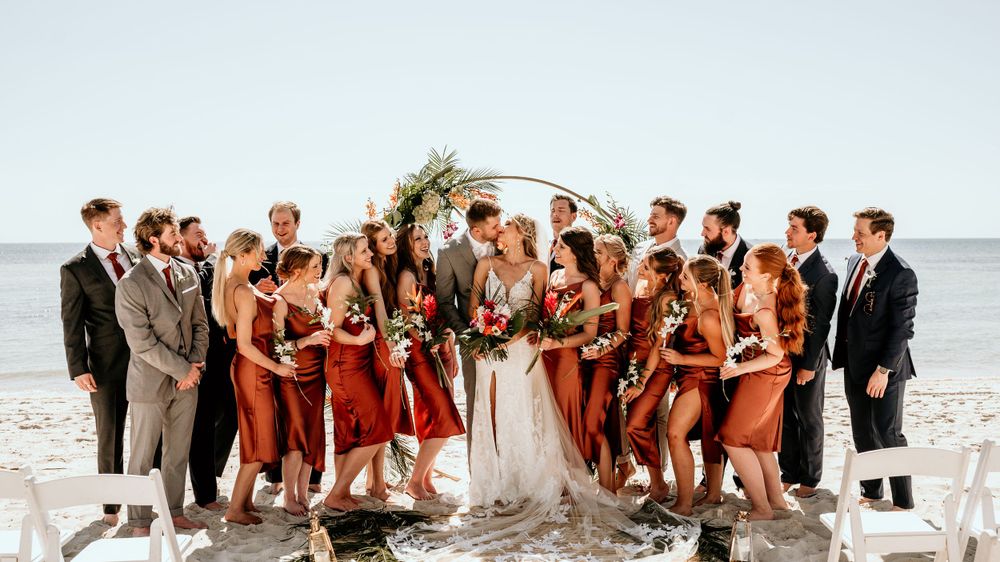 Wedding party in rust and gray attire posing together on beach.