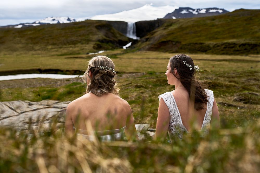 Two people sitting on a hillside overlooking a waterfall in a scenic mountain landscape.