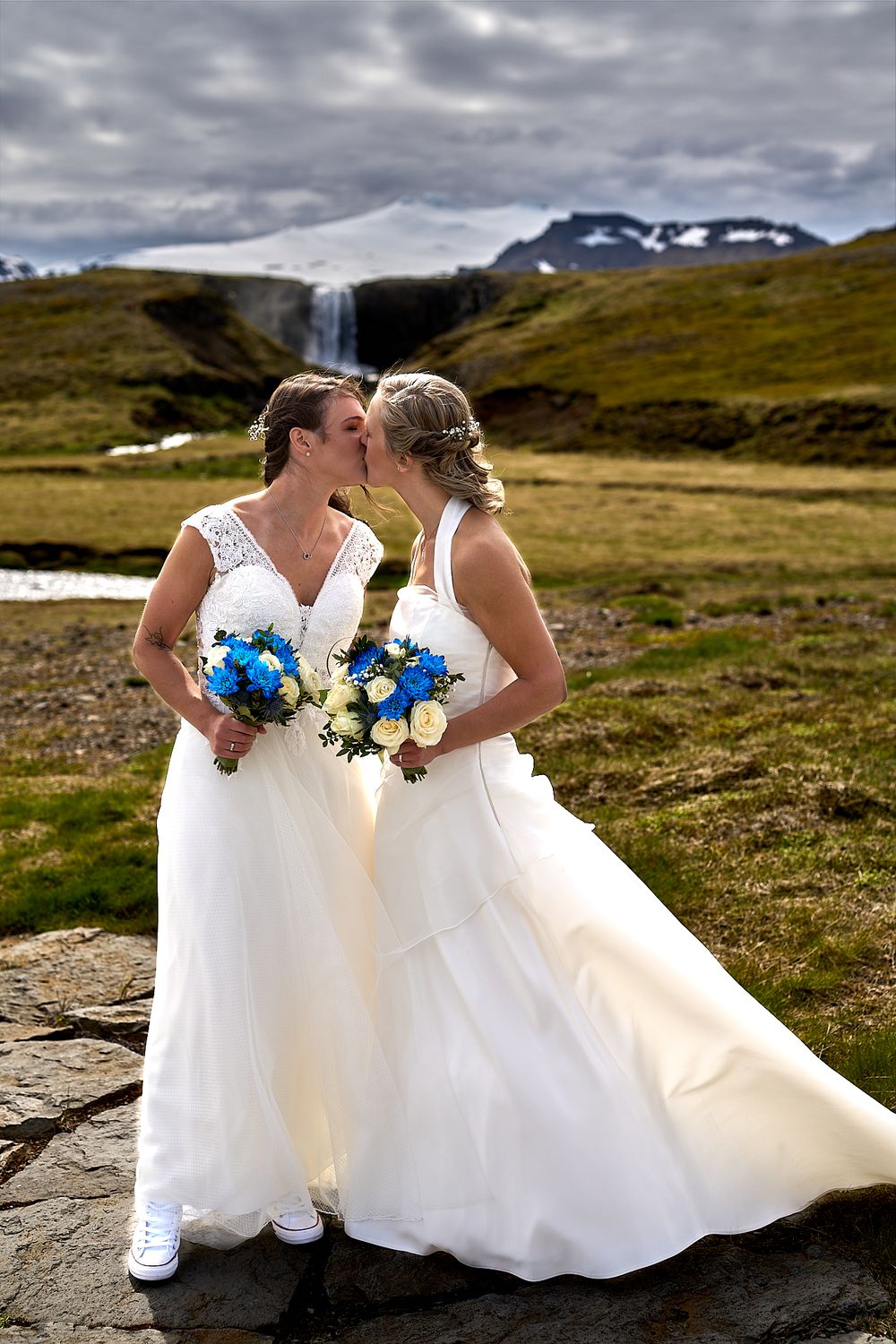 Two brides in elegant white gowns hold blue bouquets while posing in front of snow-capped mountains.