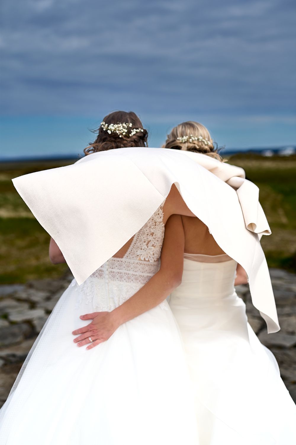 Two brides in white wedding dresses share a moment under cloudy skies in a rugged outdoor landscape.